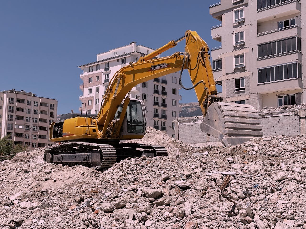 Yellow excavator working on urban construction site amidst rubble and buildings in Kahramanmaraş, Türkiye.